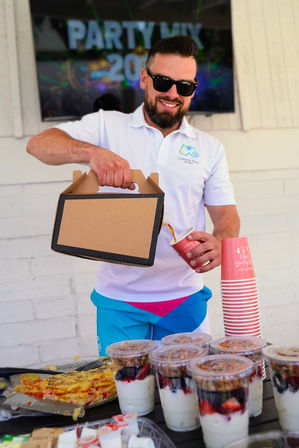Smiling server in sunglasses pours coffee from a cardboard carrier into a paper cup at an outdoor party booth with stacked cups, yogurt parfaits and pastries on the table.