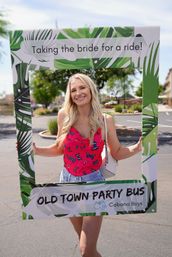 Smiling blonde woman posing in a tropical leaf photo frame that reads "Taking the bride for a ride!", wearing a pink butterfly top and denim shorts in a sunny suburban parking lot — bachelorette celebration.