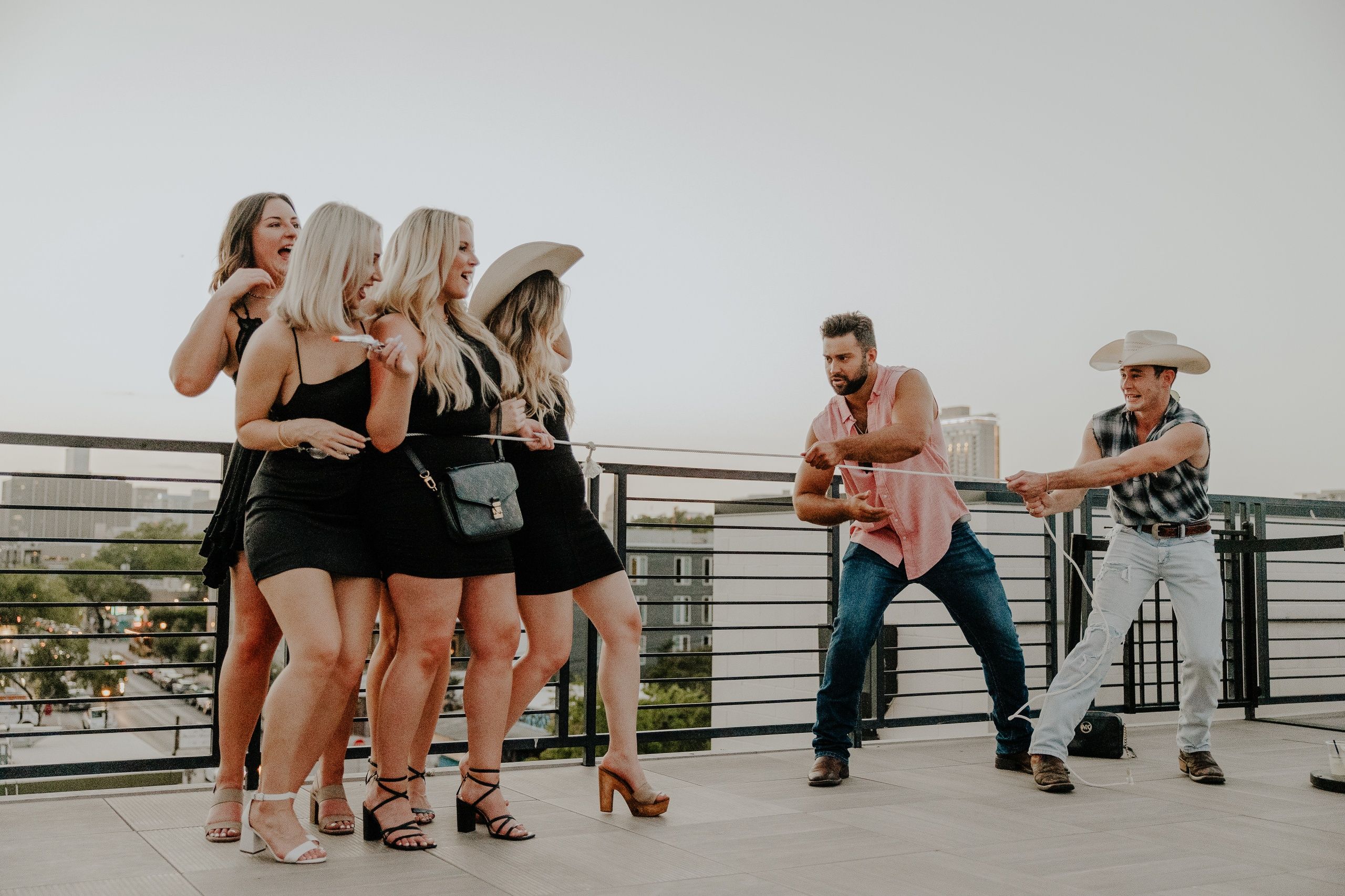Group of women in black cocktail dresses laughing on an urban rooftop terrace as two men, one in a cowboy hat, playfully tug on a rope with a city skyline at dusk.