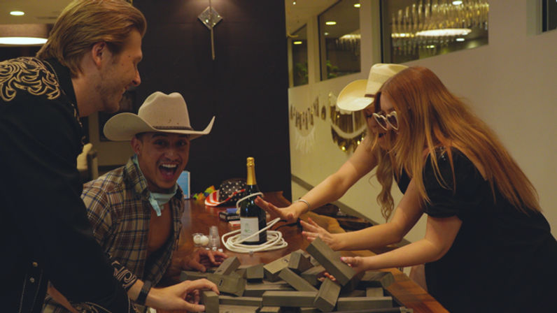 Friends playing a jumbo wooden-block game around a modern kitchen island at an indoor house party, wearing cowboy hats and playful sunglasses with champagne and festive decor on the counter.