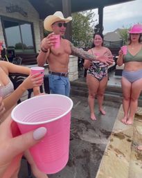 Backyard pool party scene: shirtless man in a cowboy hat and sunglasses raises a pink cup while friends in swimsuits — one wearing a pink cowgirl hat — toast on a stone patio beside the pool.