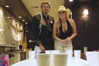 Two people in Western attire—man in embroidered cowboy shirt and woman in a white cowboy hat and black top—leaning over a table of white cups playing beer pong at an indoor party in a modern kitchen with gold decorations.