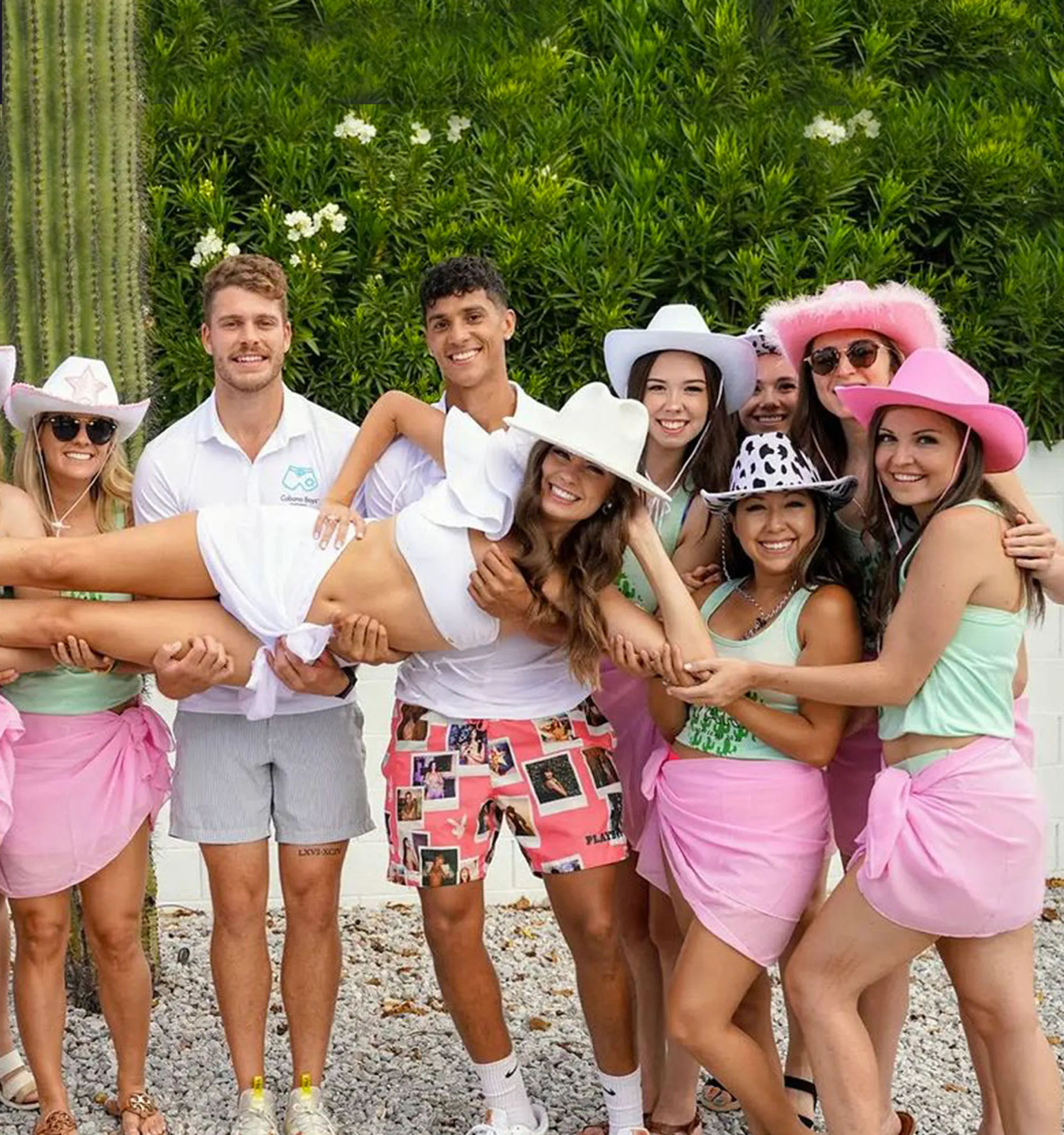 Smiling friends at a sunny outdoor party hold a woman in a white dress horizontally; group wearing cowboy hats and pink sarongs in front of cactus and green hedge.