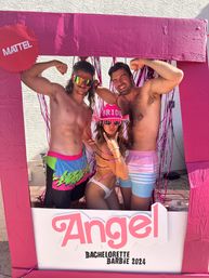 Three friends posing in a bright pink, doll‑inspired photo frame at an outdoor bachelorette party — bride in a pink 'BRIDE' cowboy hat and white bikini bottoms making a rock hand sign, flanked by two shirtless men flexing against a pink tinsel backdrop with 'Angel Bachelorette 2024' signage.