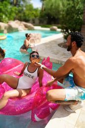 Bride in a white "BRIDE" swimsuit lounging on a pink inflatable flamingo in a backyard pool as a man feeds her a slice of pizza, sunny bachelorette pool party vibe