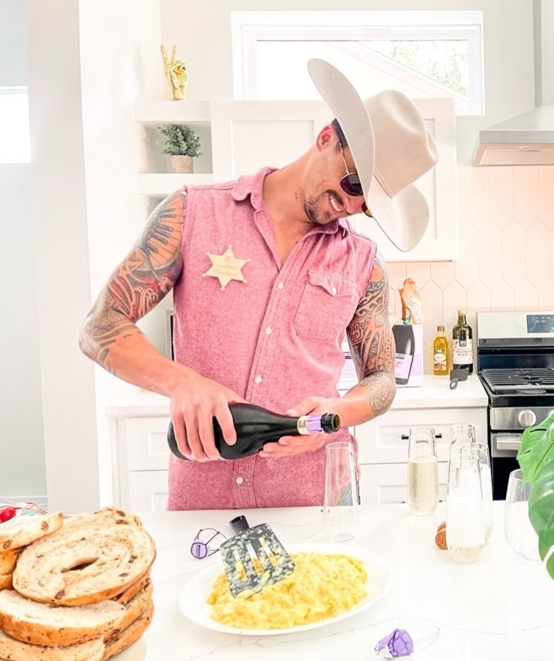 Tattooed man in a cowboy hat and sleeveless pink shirt with a sheriff star pouring sparkling wine into flutes in a bright modern white kitchen at brunch, with scrambled eggs on a plate, stacked bagels, and a stove and olive oil bottles in the background.