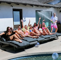 Cheerful group of women in swimsuits lounging on poolside sunbeds on a sunny backyard patio while a man sprays champagne and water splashes during a lively pool party.