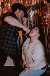 Man in a cowboy hat pouring champagne into a friend’s mouth at an indoor party photo booth with rose-gold fringe backdrop