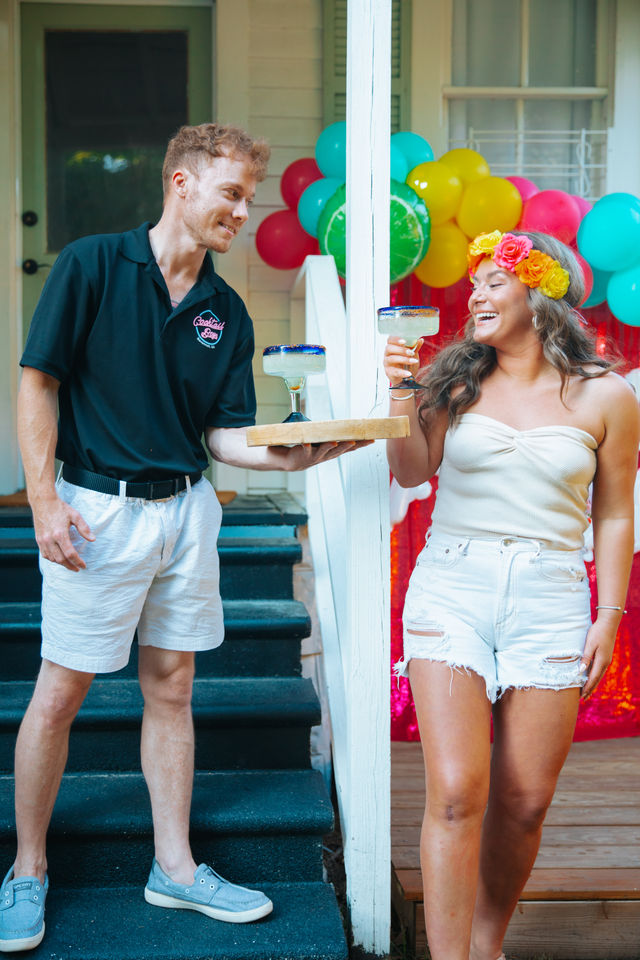 Server handing margaritas to a smiling woman wearing a flower crown on a decorated backyard porch with colorful balloons at a summer party