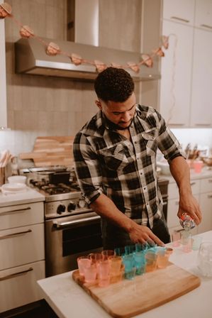 Person in a plaid shirt pouring spirits into colorful shot glasses arranged on a wooden board in a modern home kitchen with stainless steel stove and heart garland overhead