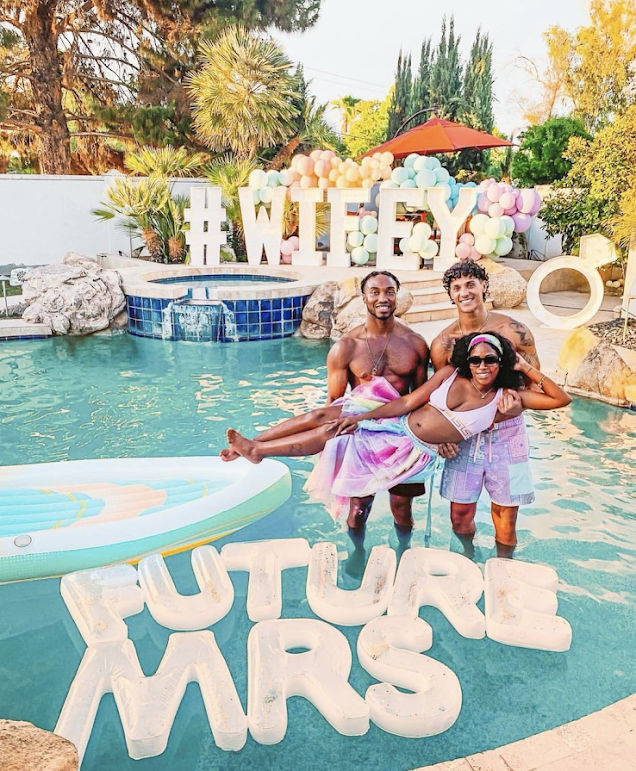 Three people posing in a sunny backyard pool at a pastel-themed engagement celebration — two standing in the water holding a smiling bride-to-be in colorful outfit, floating letters reading FUTURE MRS and a large #WIFEY balloon display with palm trees and a pool float nearby.
