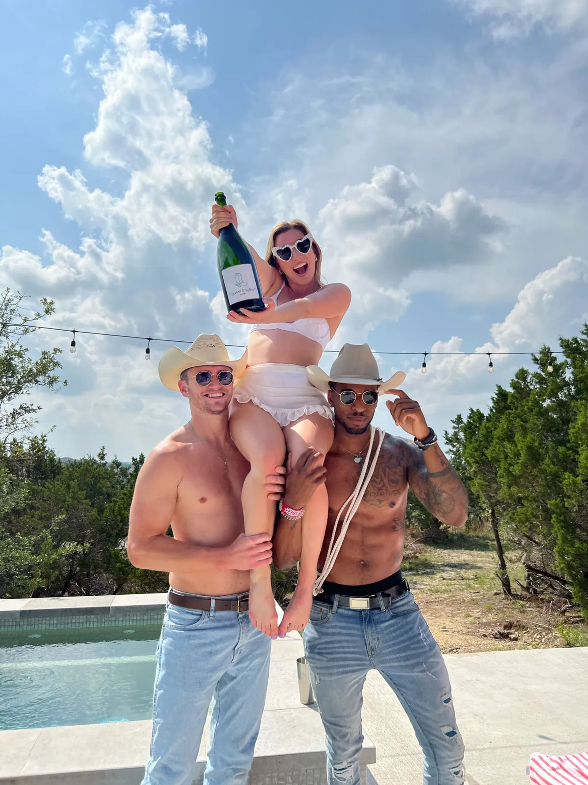 Three friends at a sunny outdoor pool party: two shirtless men in cowboy hats lift a woman in a white bikini who holds a large champagne bottle, with blue sky, string lights, and trees in the background.