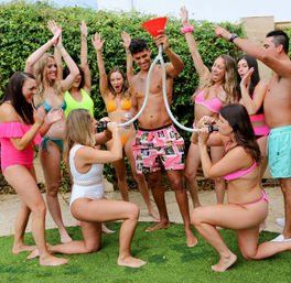 Sunny backyard summer party with friends in colorful swimsuits; two women kneel using a funnel beer-bong held by a smiling man while the group cheers.