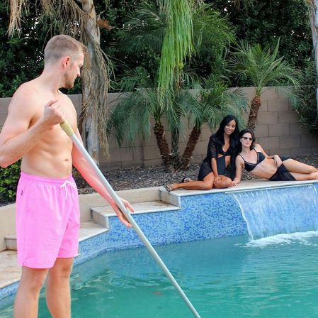 Shirtless man in bright pink swim trunks skimming a backyard pool with a long pole while two women in black swimwear lounge on the tiled edge near a small waterfall and palm trees.