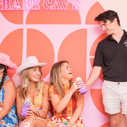 Group of friends laughing in front of a retro pink-and-orange mural with a neon sign, women in bright summer dresses and a cowboy hat holding canned drinks while a man reaches out
