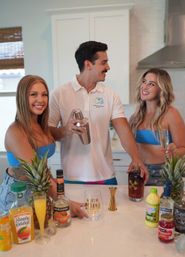 Three friends making tropical cocktails at a modern white kitchen island — bartender shaking a cocktail shaker while two women in blue tops smile amid pineapples, orange juice, triple sec and rum for a summer home-bar vibe.