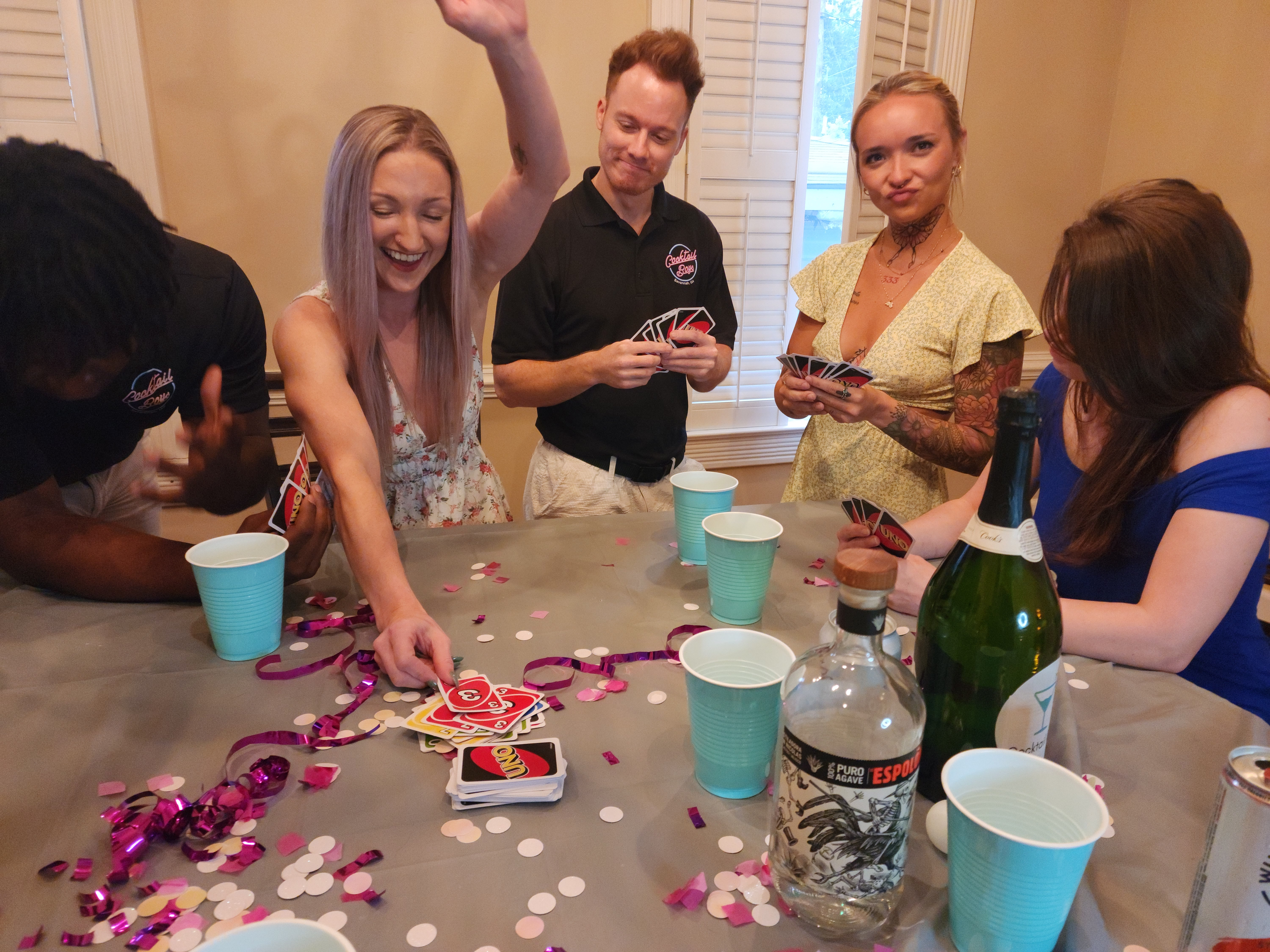 Five friends gathered around a table at an indoor house party, laughing and playing UNO amid confetti, plastic cups and bottles.