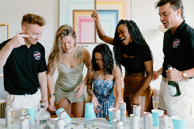 Five friends laughing and cheering around a living-room table playing a cup-flip game, surrounded by blue plastic cups, canned drinks, a bottle of sparkling wine and confetti at an indoor celebration.