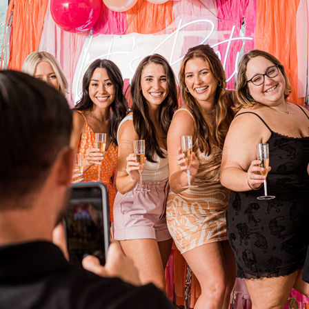 Five smiling women posing with champagne flutes at a colorful pink-and-orange party photo booth with balloons and streamers, phone in foreground capturing the girls' night celebration.