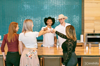Group of five adults toasting with small cups across a wooden kitchen island in a bright modern space with teal tile backsplash; two men wearing cowboy hats smile behind the counter.