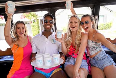 Four friends in colorful summer outfits cheer with white cups inside a party trolley—host holds a tray of drinks as they smile on a sunny suburban street.