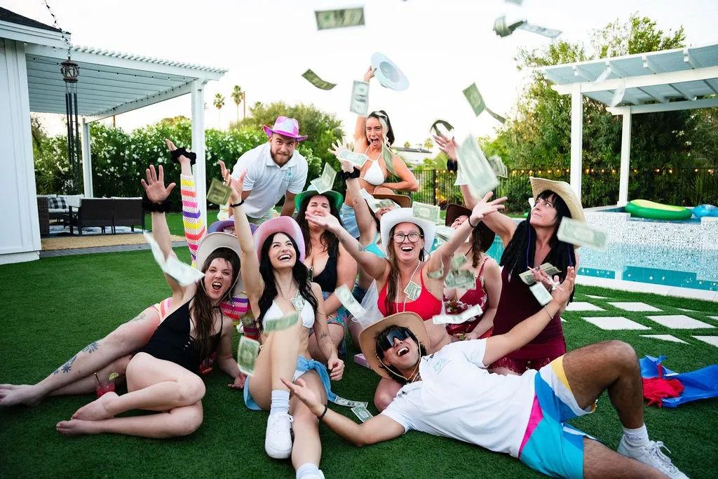 Sunny backyard pool party on artificial turf: group of friends in swimsuits and cowboy hats laughing and tossing dollar bills near a pool and pergola.