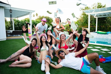 Friends at a sunny backyard pool party on artificial turf, wearing swimsuits and cowboy hats, laughing and tossing dollar bills in the air near a pool and pergola.