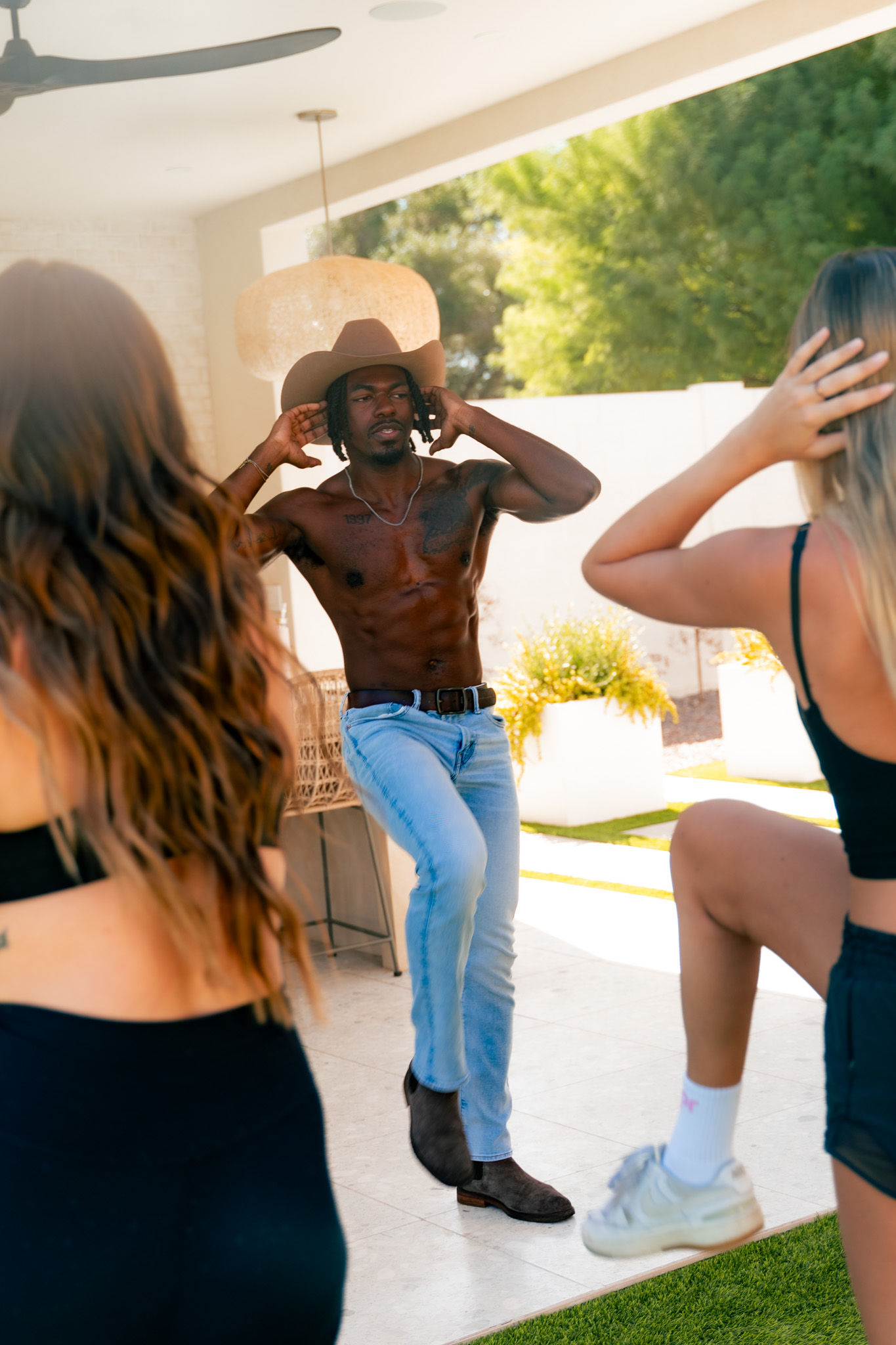 Shirtless man in a cowboy hat, jeans and boots leading a playful dance on a sunny backyard patio while two friends mimic him.