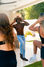 Shirtless man in a cowboy hat, jeans and boots leading a playful dance on a sunny backyard patio while two friends mimic him.