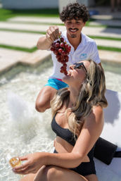Poolside: smiling man feeds a bunch of red grapes to a woman in a black swimsuit lounging by a sunlit pool with water jets, sunglasses and a cocktail.