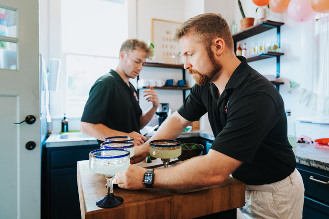 Two bartenders in black shirts preparing margarita-style cocktails in blue-rimmed glasses with lime wedges at a bright neighborhood kitchen bar counter