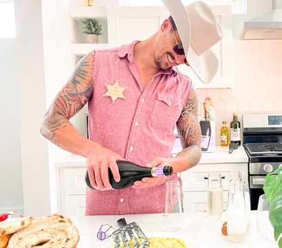 Smiling tattooed man in a cowboy hat and sleeveless pink shirt with a novelty sheriff star pours sparkling wine into flutes on a bright modern kitchen island with scrambled eggs, stacked bagels and breakfast items.