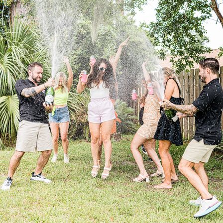 Six friends at a backyard summer party popping champagne and cheering as spray drenches them near palm plants and a wooden fence