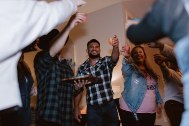 Group of friends toasting with colorful shot glasses in a living-room house party, smiling adults in casual flannel and denim with one person holding a tray of shots.