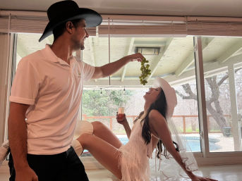 Playful scene: man in a black hat offers green grapes to a woman in a white fringe dress and veil as she leans back with a drink by a sliding glass door overlooking a backyard patio and pool.