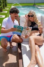 Two people poolside at a backyard pool: woman in black bikini lounges on a sunbed with sunglasses and champagne while a man in a white polo and colorful swim trunks kneels beside her reading a magazine, green lawn and water fountain in background.
