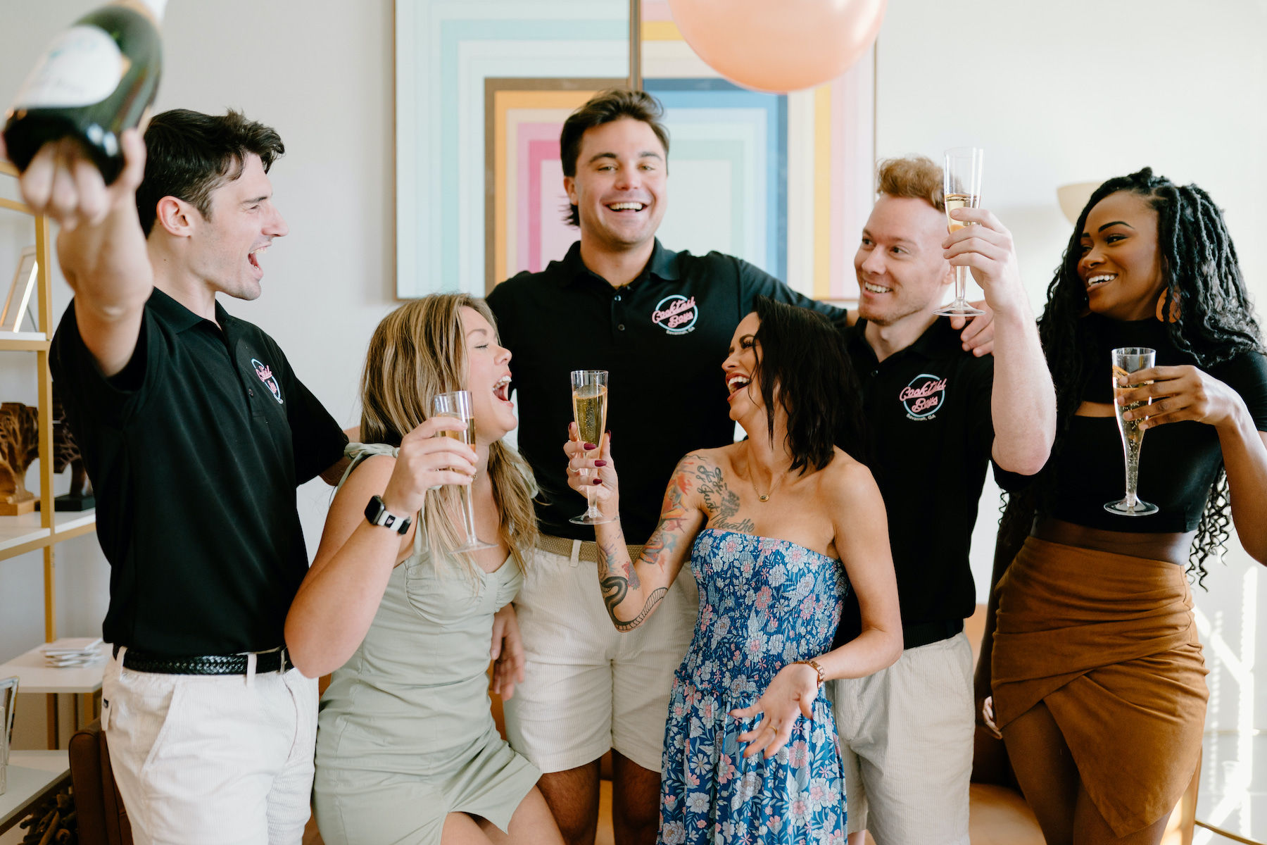 Six friends laughing and toasting with champagne flutes in a bright living-room celebration, casual summer outfits and a balloon overhead.