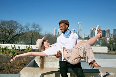 Sunny rooftop photoshoot: smiling man lifts woman in a wide-brim hat and floral skirt against a downtown skyline and clear blue sky, with urban greenery in the foreground.