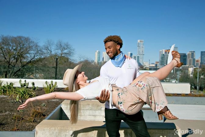Sunny rooftop photoshoot: smiling man lifts woman in a wide-brim hat and floral skirt against a downtown skyline and clear blue sky, with urban greenery in the foreground.