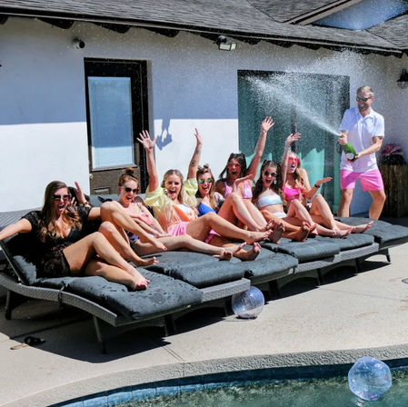 Backyard pool party with a row of women in colorful swimsuits on poolside daybeds cheering as a man sprays champagne over them on a sunny patio by a swimming pool