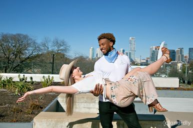 Playful scene of a man lifting a smiling woman in a sun hat and floral skirt on a rooftop with a clear blue sky and downtown skyline in the background.