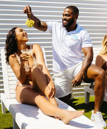 Playful poolside scene: woman in a bikini lounges on a white sunbed with a cocktail while a man in a white polo dangles a bunch of green grapes above her in bright sunlight.