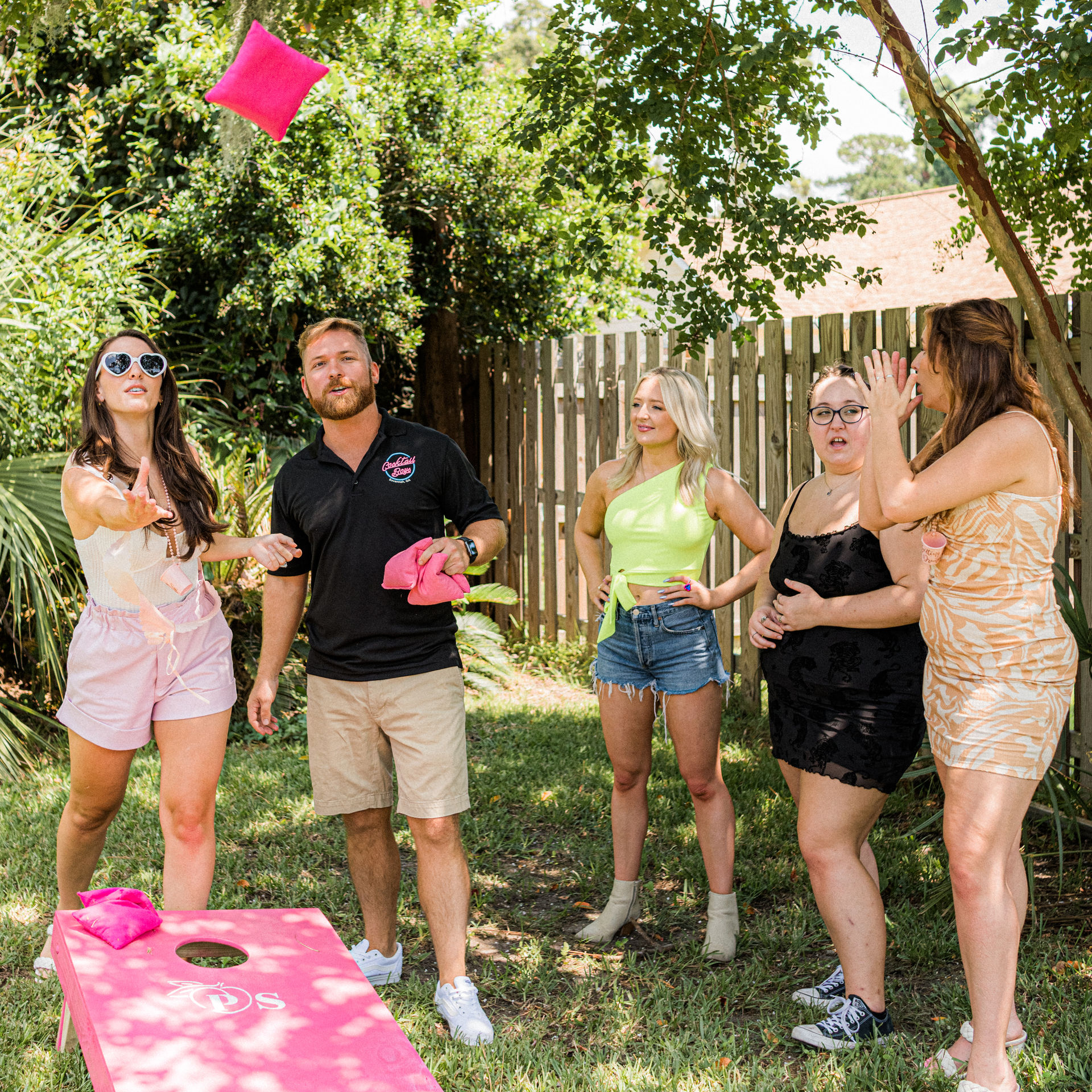 Five friends playing cornhole in a sunny backyard — a woman tosses a bright pink beanbag toward a pink cornhole board while others cheer beside a wooden fence and leafy trees, casual summer outfits.
