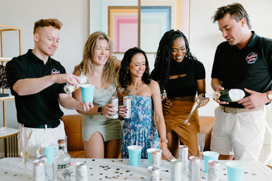 Group of five friends at an indoor living-room party pouring champagne and canned cocktails into blue cups over a confetti-covered table — bright, modern at-home celebration.