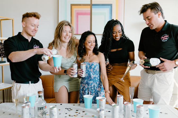 Group of five friends at an indoor living-room party pouring champagne and canned cocktails into blue cups over a confetti-covered table — bright, modern at-home celebration.