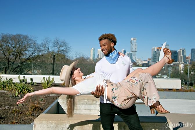 Smiling man in a white polo and blue bandana lifts a woman in a wide-brim hat, white top and floral pants on a sunny rooftop garden with a downtown skyline in the background.