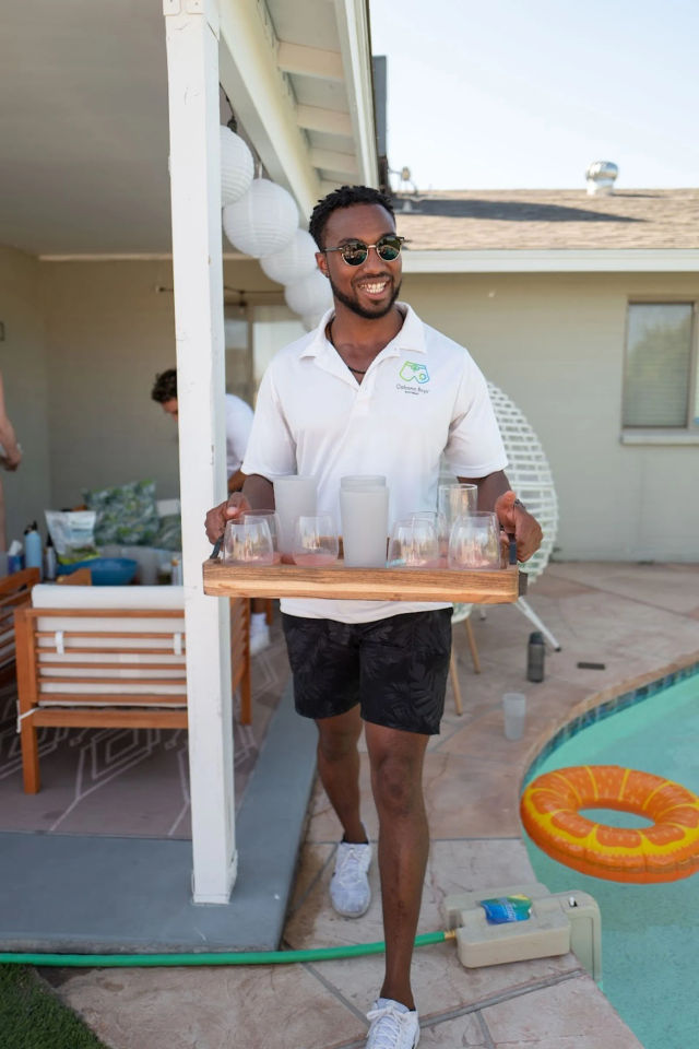 Smiling server in sunglasses carrying a wooden tray of glasses at a sunny backyard pool party, wearing a white polo and black shorts with patio seating and an orange inflatable ring visible