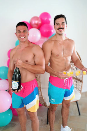 Two shirtless men in matching bright pink, teal and yellow swim shorts smile while one holds a large champagne bottle and the other a tray of orange shots, standing by a wicker chair with pink and teal balloons in the background — festive indoor summer party vibe.