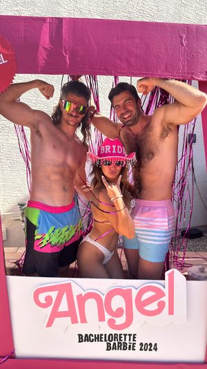 Three friends posing in a bright pink cardboard photo-frame prop at a sunny backyard bachelorette party — bride in a pink "BRIDE" fringe hat and white bikini, flanked by two shirtless men flexing with metallic tinsel backdrop and "Bachelorette 2024" sign.