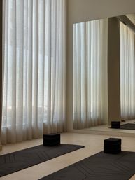 Minimal, light-filled urban yoga studio interior with black mats and stacked foam blocks on a pale floor, floor-to-ceiling sheer curtains and a large mirror reflecting soft natural light.